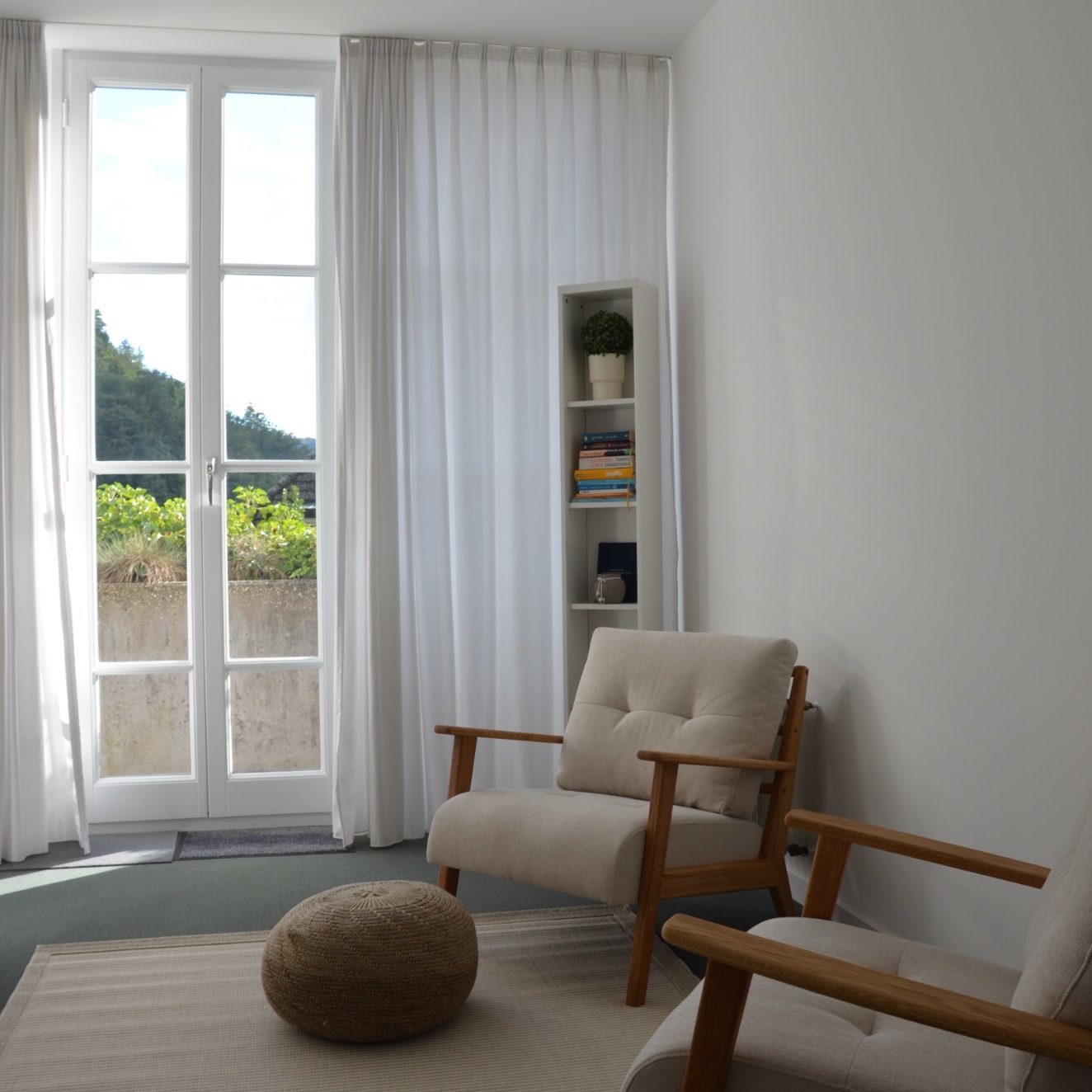 Heller Raum mit zwei Sesseln, einem Teppich und einem Regal, große Fenster mit Gardinen.