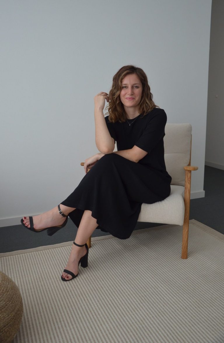 Frau in schwarzem Kleid sitzt entspannt auf einem Stuhl in einem hellen Raum.